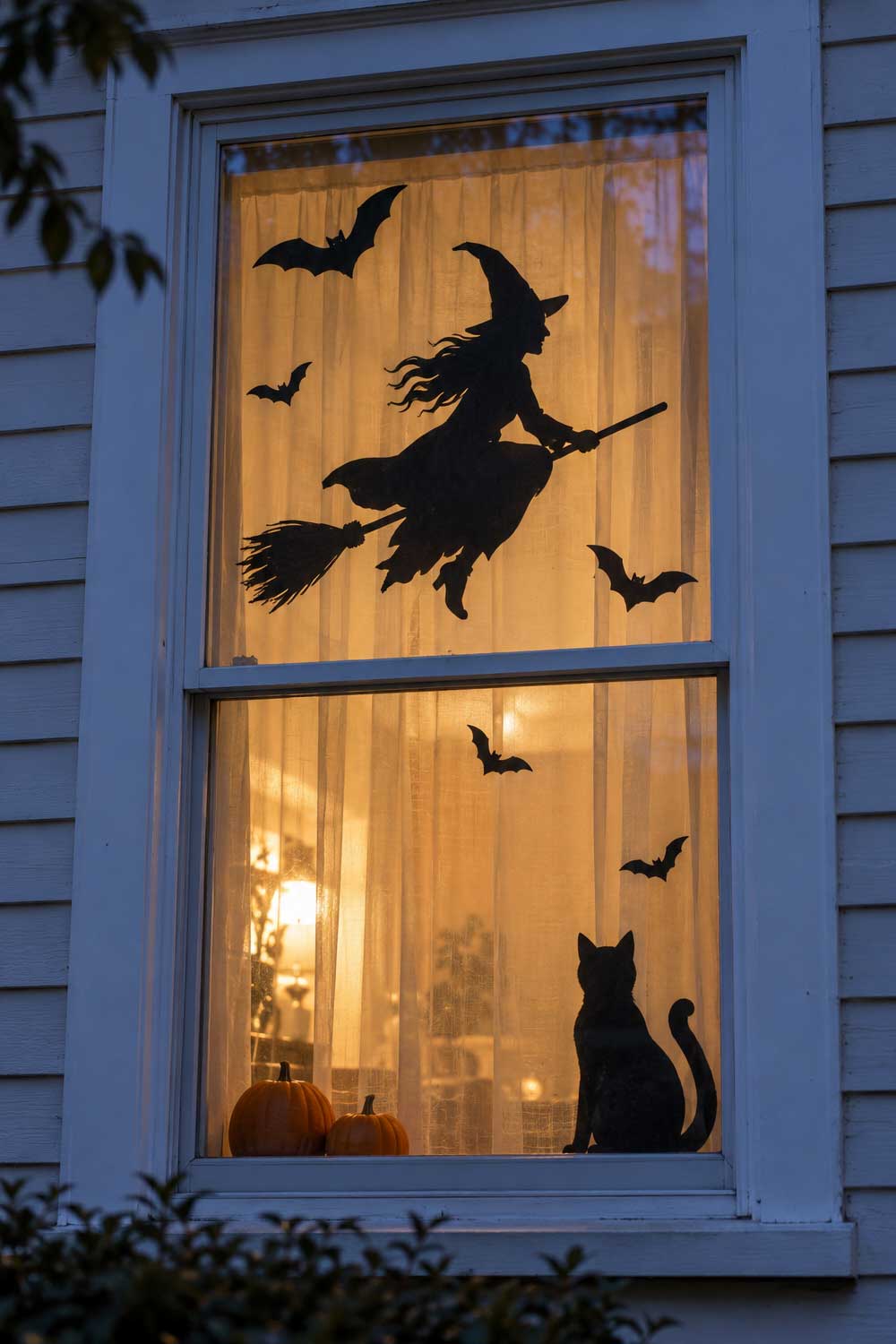 Halloween window with black witch, cat, and bat silhouettes against warmly lit sheer curtains at dusk